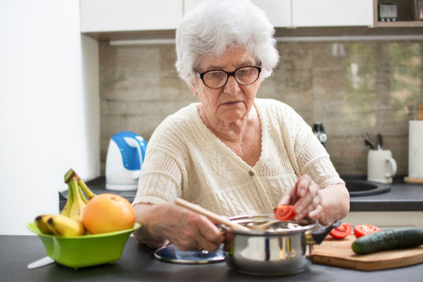 A senior woman with white hair and black rimmed glasses stands at the kitchen counter as she puts a tomato into a pot. There is a green bowl of fruit next to her.