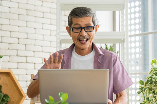 A man with a white t-shirt and purple short-sleeved shirt smiles at his waves at someone on his computer. 