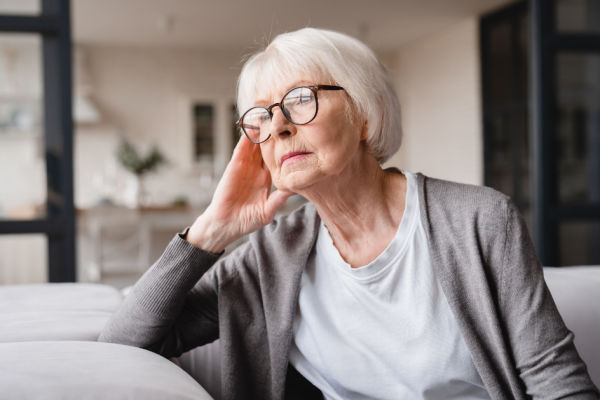 An elderly woman with white hair, glasses, a white shirt, and grey sweater looks off to the side. She is deep in thought as she rests her right hand on her face. 