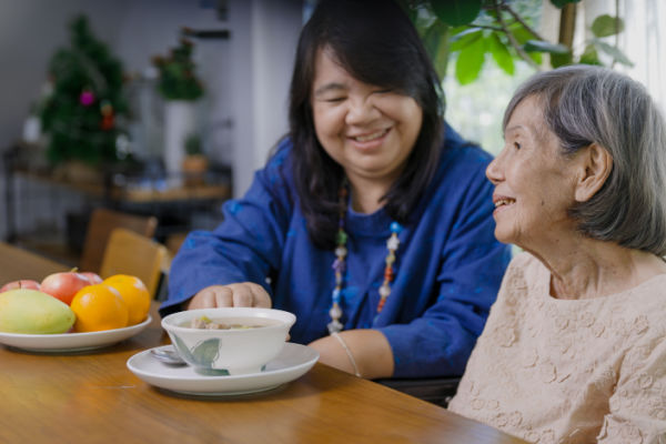 A younger woman sits at a table with an older woman. They are both smiling, and there is some food on the table such as a bowl of fruit. 