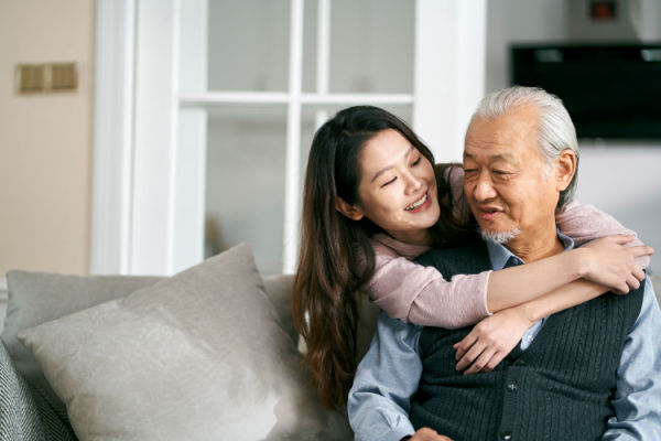 A young woman puts her arms around an elderly man's neck and shoulders from behind as she lovingly looks at him.