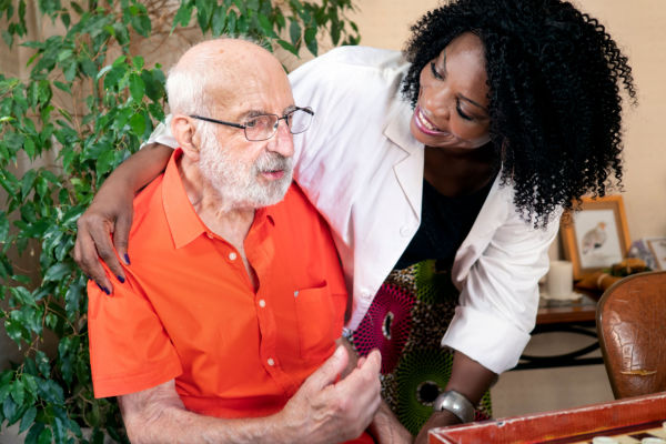 An elderly man with a white beard, glasses, and an orange shirt sits as a nurse or home health aide leans over him and smiles, with her arm around him.