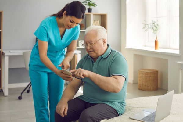 An older man sits on a bit as a home health aide or nurse stands beside him. She is showing him something on her phone as he uses his finger to point to it or scroll 