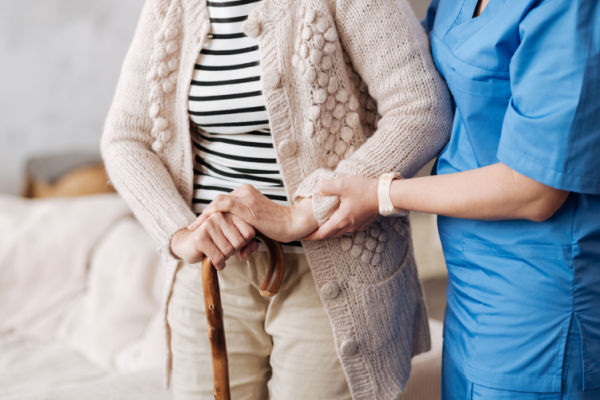 A nurse or home health aide assists an elderly woman as she walks with a cane. 
