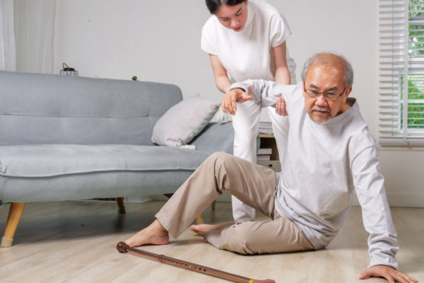 An older man sits on the floor with his cane in front of him. He appears to have fallen, as a younger woman has her hand on his arm to help him up.