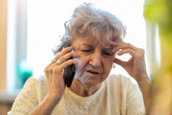 An older woman talks on the phone with a look of confusion on her face. One hand holds the phone while the other hand gently touches her forehead. 