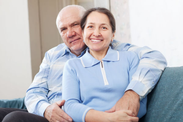 A smiling senior man and woman sit on a couch. The woman is leaned back against the man, and his arm is around her.