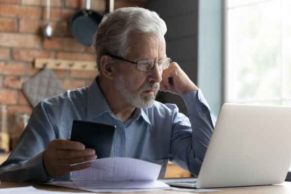 A man with grey hair and glasses stares worriedly at a computer as he holds a calculator in his hand. 