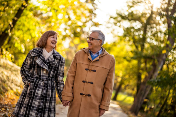 A senior couple smiles as they hold hands and walk outside on a brisk day. 