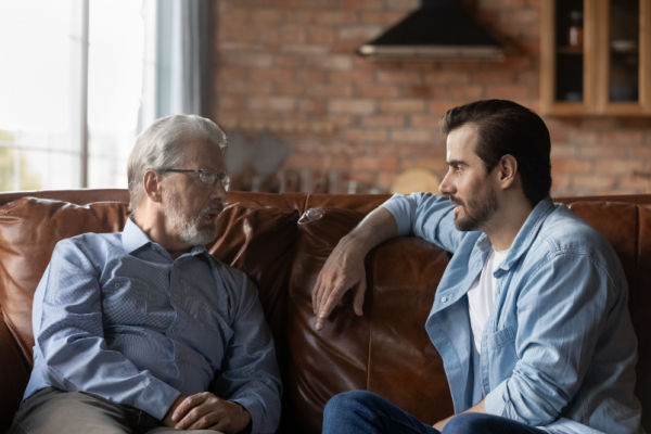 A father and adult son have a conversation while sitting on a brown leather couch. 