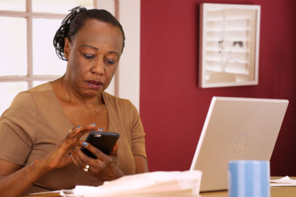 A woman sits in front of a laptop and looks down at a cellphone she is holding. She has a serious look of concentration on her face. 