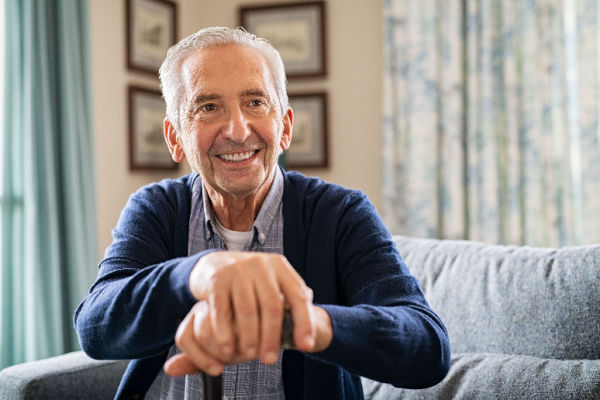 A smiling older man sits on a couch as he rests his hands on his cane. 