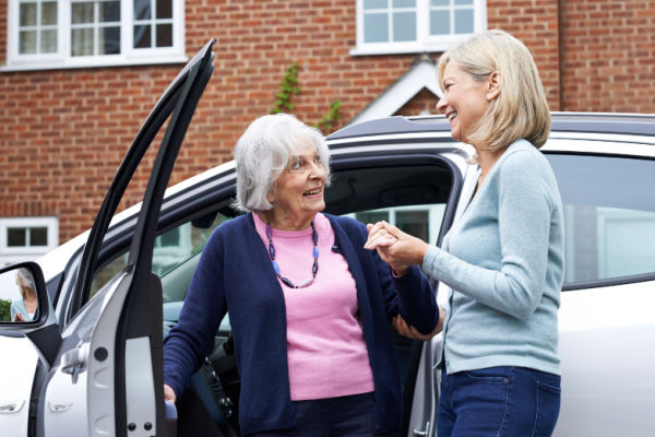 A senior woman is helped out of the car by a middle-aged woman. Both women are smiling.