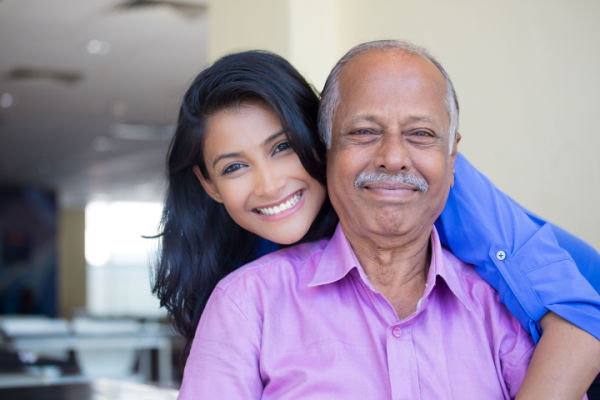 A young woman stands behind a senior man. She has one arm around him from behind, and they are both smiling.