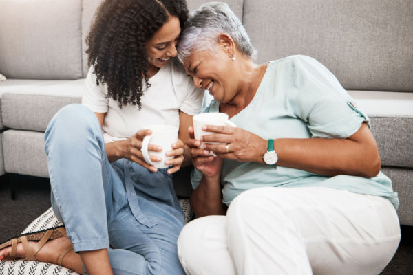 A young woman and a senior woman sit on the floor with their backs up against the couch. Their heads are touching as they smile and laugh. They are both holding mugs in their hands. 