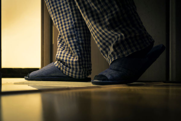A senior in checkered pajamas and grey slippers walks out of a dark room into the hallway. 