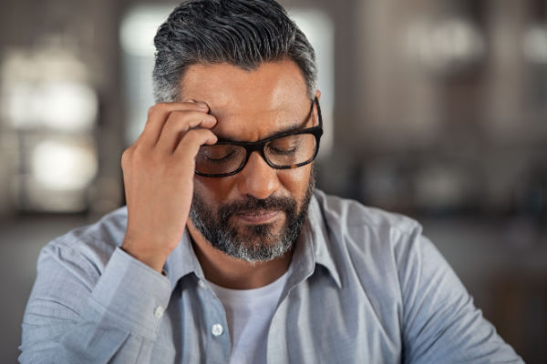 A man with dark grey hair and a mustache and beard closes his eyes as he touches his fingers to his forehead. He looks tired and stressed. 