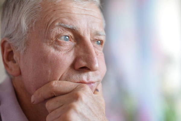 An older man with sad, pensive eyes holds his hands to his chin and mouth and he looks off to the side.