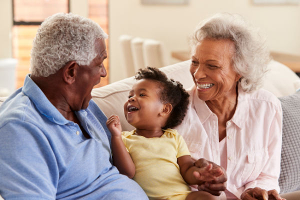 A senior couple sits on a couch as they smile and laugh with their toddler grandson.