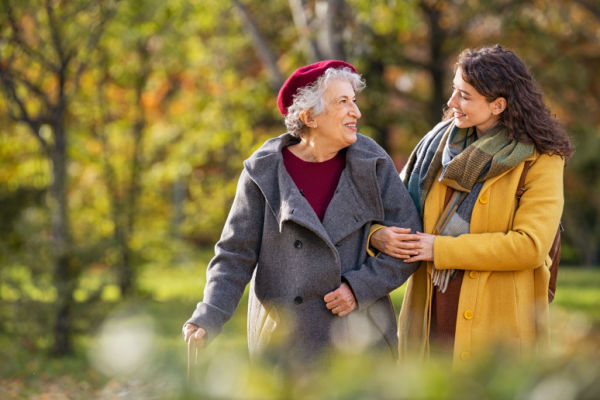 A young woman and a senior woman dressed in winter gear link arms as they walk together outside. 