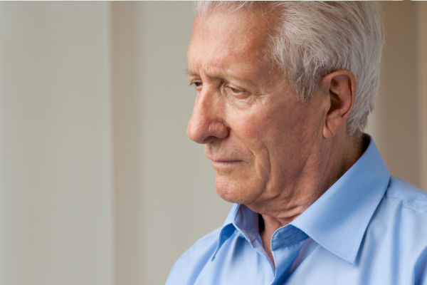 A senior man in a blue buttondown shirt pensively looks to the ground. He has an expression of worry and sadness on his face.