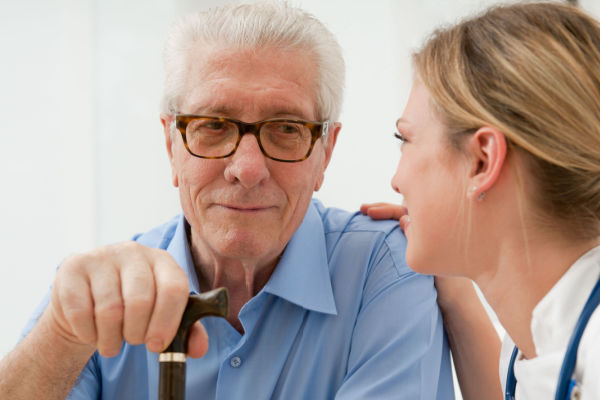 A senior man with glasses smiles slightly as he looks at a female doctor or nurse sitting next to him. The nurse has his hand on the man's shoulder. 