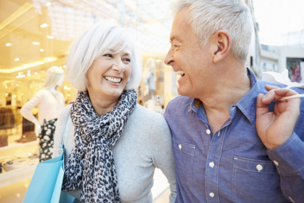 A senior man and woman smile at eac other as they walk inside at a mall.