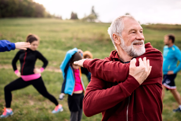 A senior man in a sweatshirt smiles as he stretches outside, along with other people. 