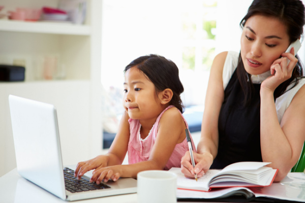 A mother is on the phone as she takes notes in a notepad. A child is next to her on a laptop computer.