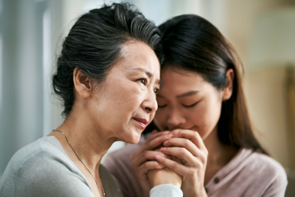 An adult daughter kisses her mother's hand as the mother stares off into the distance.