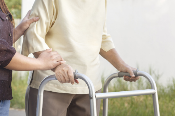 A senior woman uses a walker while supported by a loved one or caregiver.