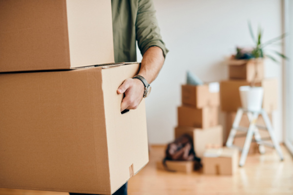 An adult carries moving boxes while some boxes sit on the floor in the background.