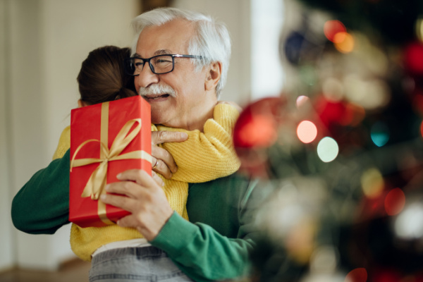 A senior man hugs a child as he holds a holiday gift next to a Christmas tree.