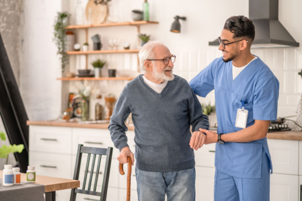 A caregiver gently helps an older man while in the kitchen, as they both smile. 