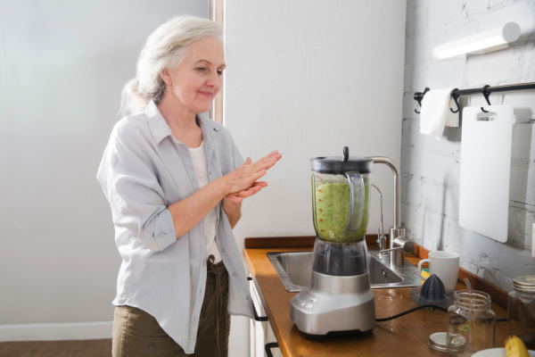 A senior woman stands in the kitchen and prepares a green smoothie. 