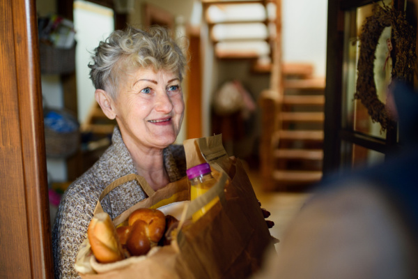 A senior woman smiles at her front door as she receives a grocery delivery. 