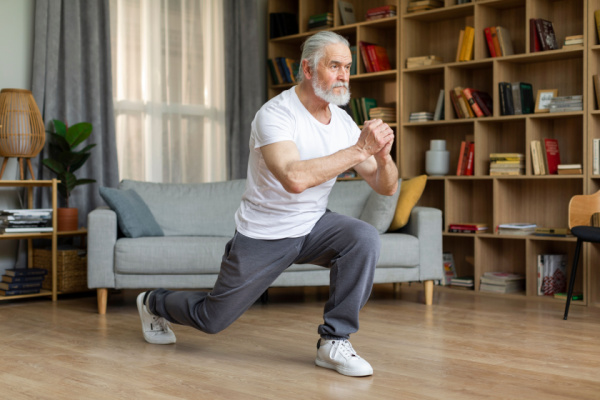 A senior man in a white t-shirt and grey sweat pants does a lunge. 