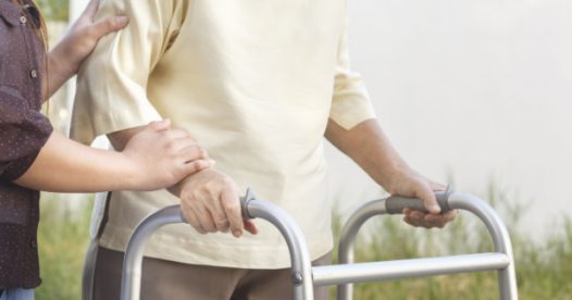 A senior woman uses a walker while supported by a loved one or caregiver.