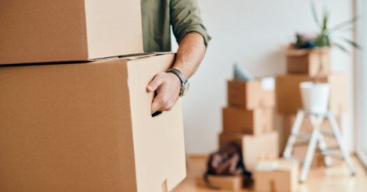 An adult carries moving boxes while some boxes sit on the floor in the background.