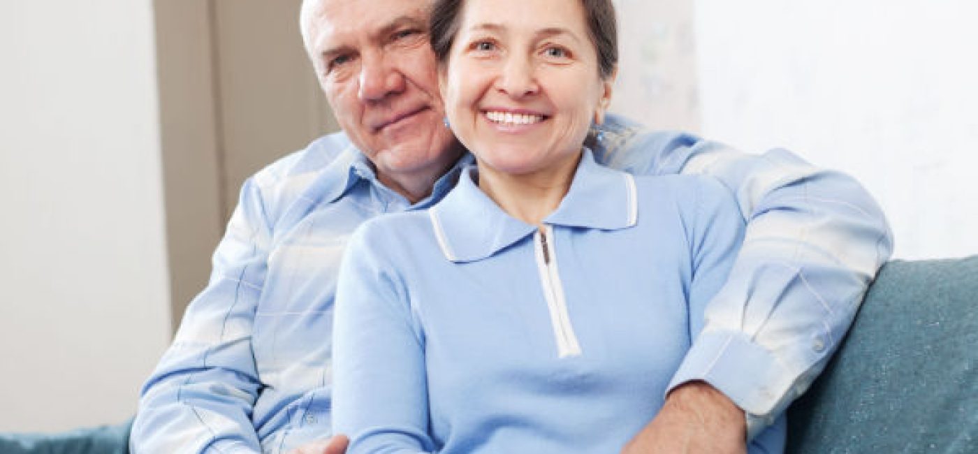 A smiling senior man and woman sit on a couch. The woman is leaned back against the man, and his arm is around her.