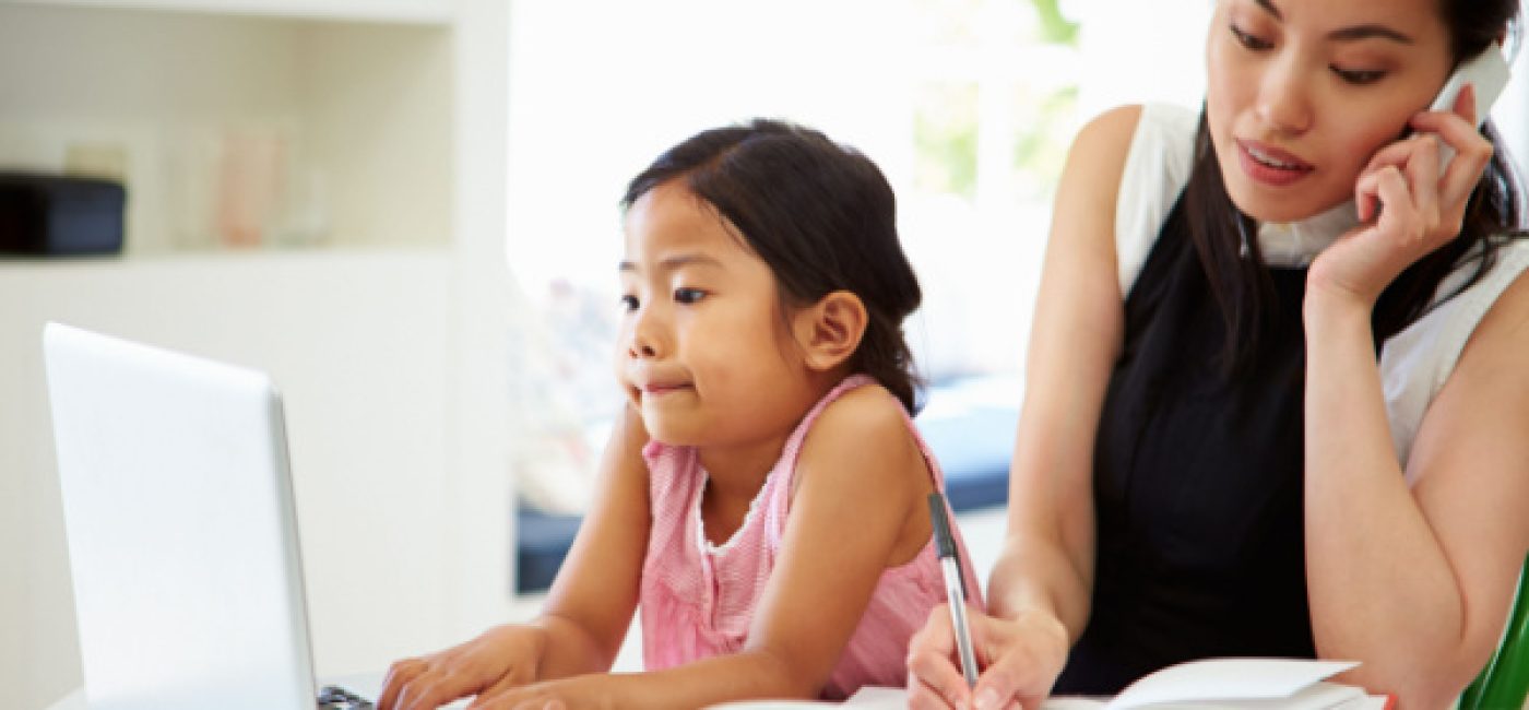 A mother is on the phone as she takes notes in a notepad. A child is next to her on a laptop computer.