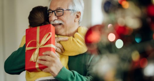 A senior man hugs a child as he holds a holiday gift next to a Christmas tree.