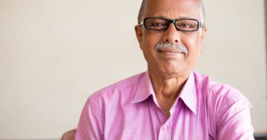 A senior man in glasses and a pink polo shirt smiles at the camera.