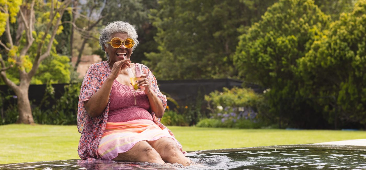 A senior woman smiles while she sips on a drink with her feet in the water.