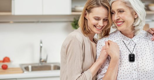 A senior woman and younger woman look at each other and smile as they stand in the kitchen.